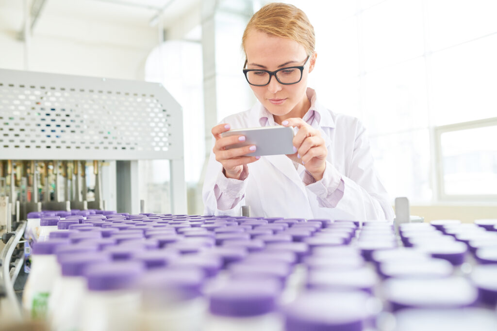 Beautiful inspector wearing eyeglasses and white coat taking picture on modern smartphone while checking quality of soy milk at production department of dairy plant