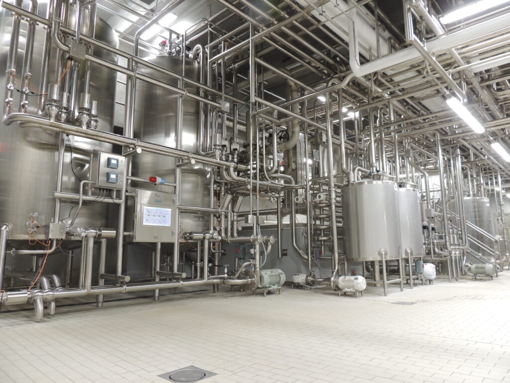 An image showcasing meticulously installed stainless steel hygienic piping systems and tanks by JBT A&amp;amp;B Process Systems, within a food and beverage production facility. 